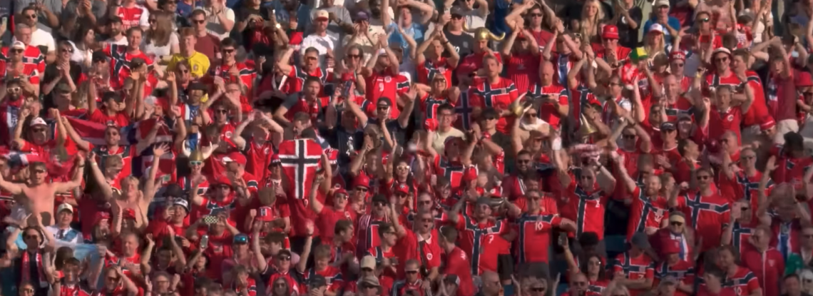 Norwegian supporters in red national team shirts cheer and wave flags from packed stadium stands during a World Cup match.