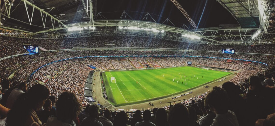 Wide-angle view of a packed football stadium at night, with floodlights, spectators filling the stands, and players on the pitch.