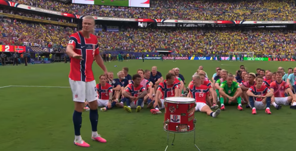 Norway’s football team sits on the pitch after beating Brazil, while one player stands beside a red drum in front of a packed stadium.